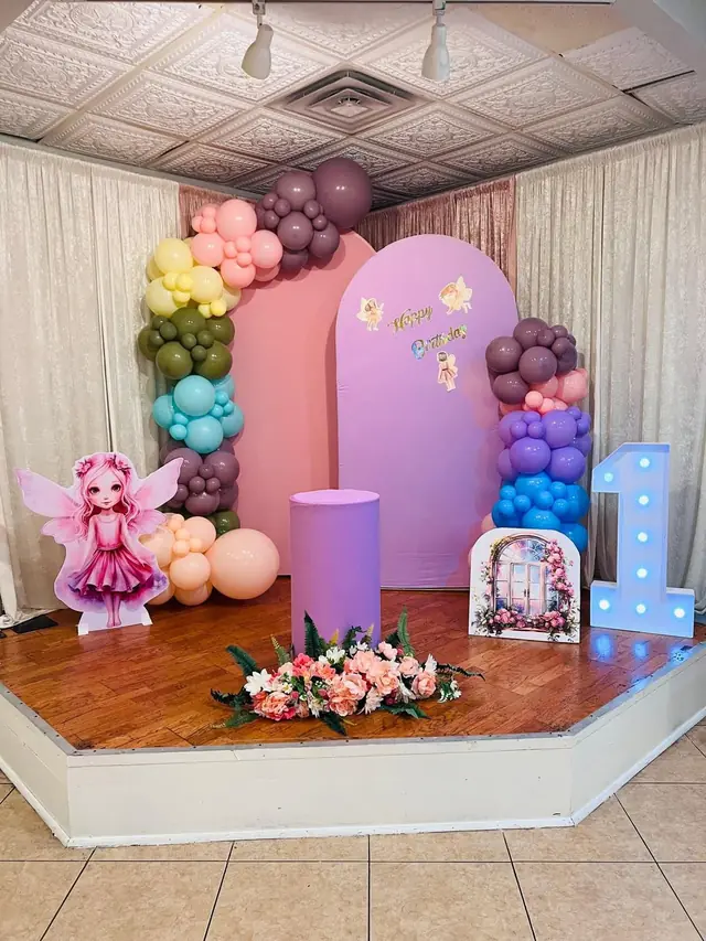 Fairy themed first birthday stage with a pastel balloon garland, pink and lilac arch panels and a light-up number one